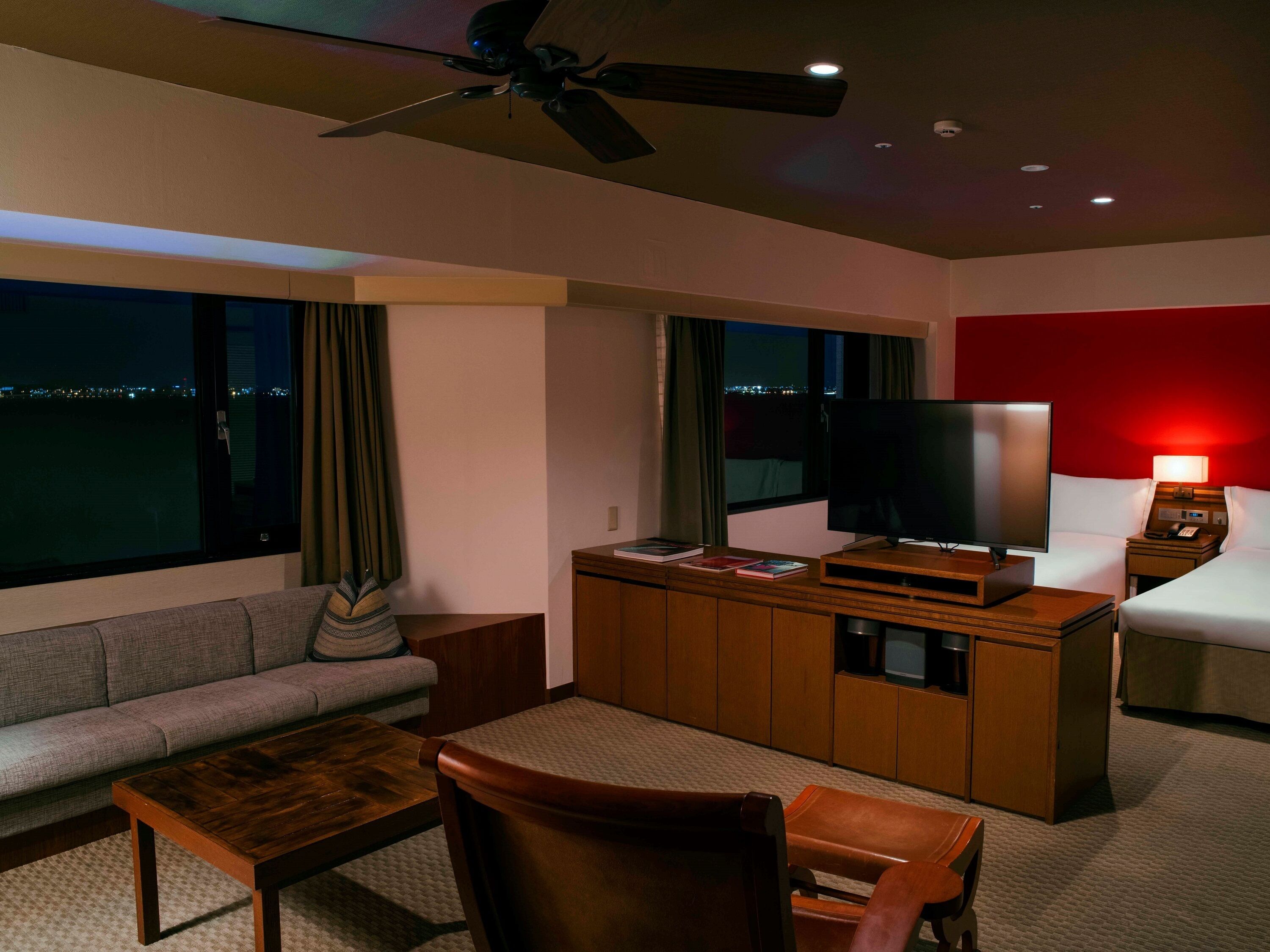Habitación de hotel con sofá, una mesita de centro de madera, televisor sobre un mueble de madera y cama frente a una pared con detalles en rojo, además de un ventilador de techo.