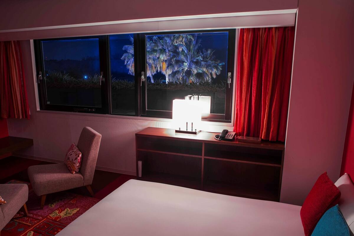 A hotel room with a large bed, a desk with a lamp, a chair, and a view of palm trees through the windows.