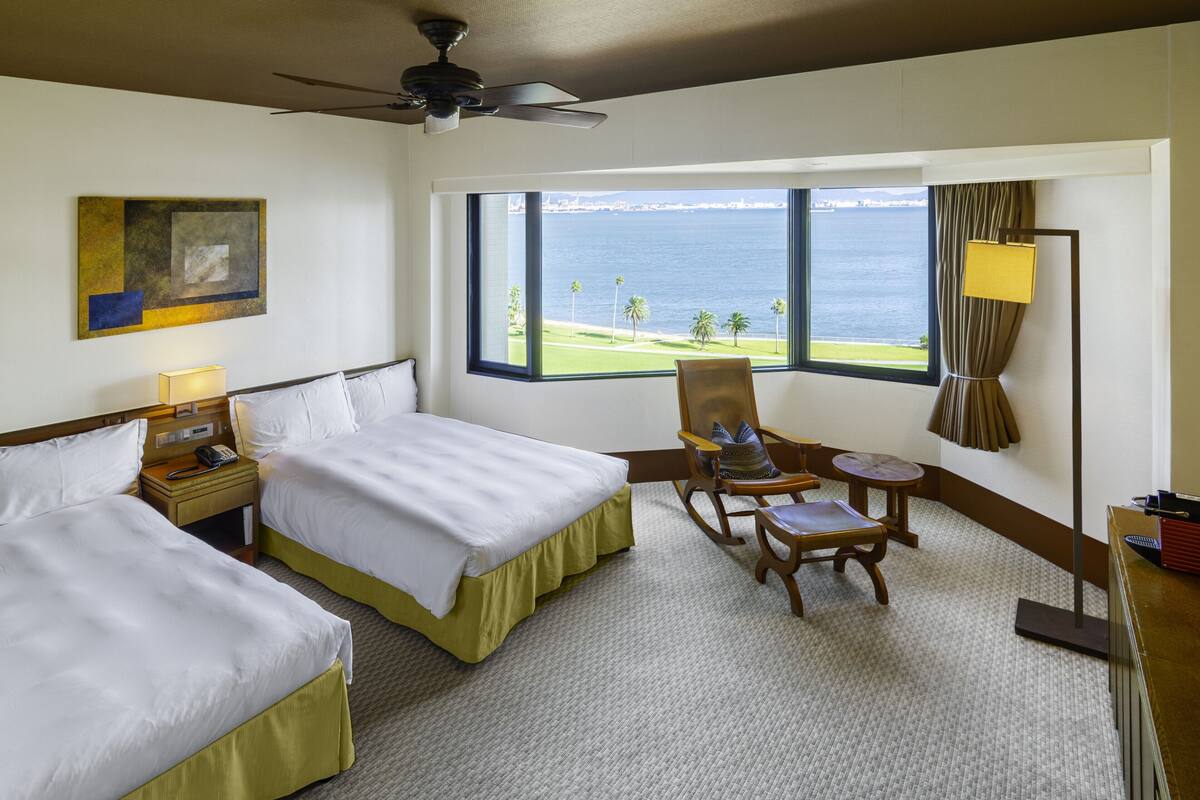 A hotel room with two beds, a chair, a desk, a lamp, and a view of the sea.