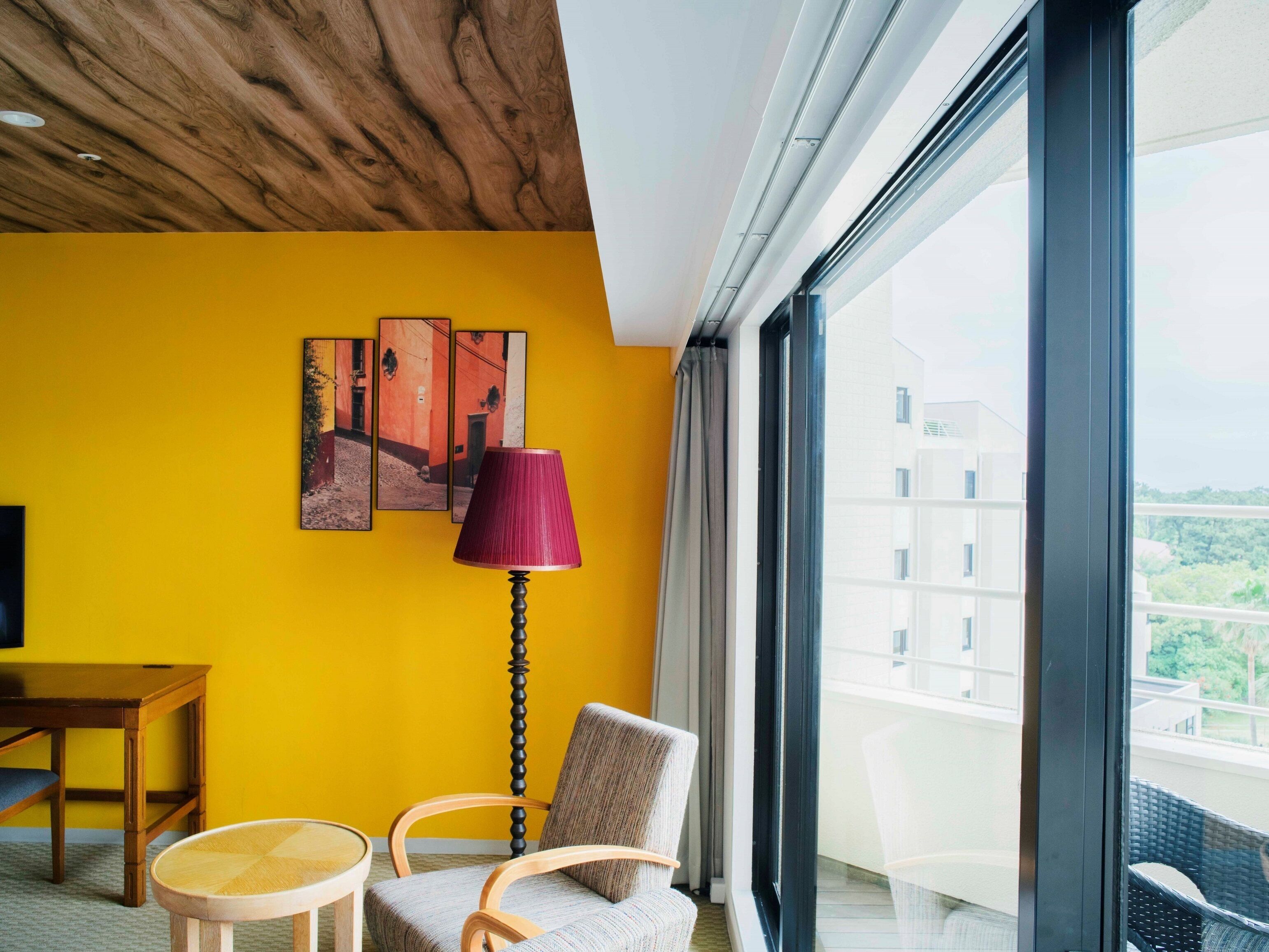A hotel room with a yellow wall, a wooden ceiling, a large window, a table, two chairs, and a lamp.