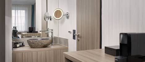 A modern bathroom with a stone basin sink, a round mirror, and a coffee machine.