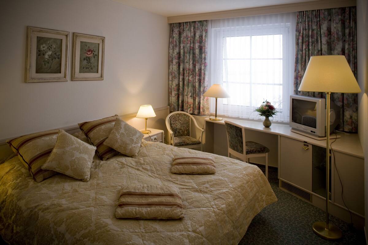 Une chambre d’hôtel avec un lit, deux lampes, une télévision, un bureau agrémenté d’un vase de fleurs et deux tableaux floraux encadrés accrochés au mur.