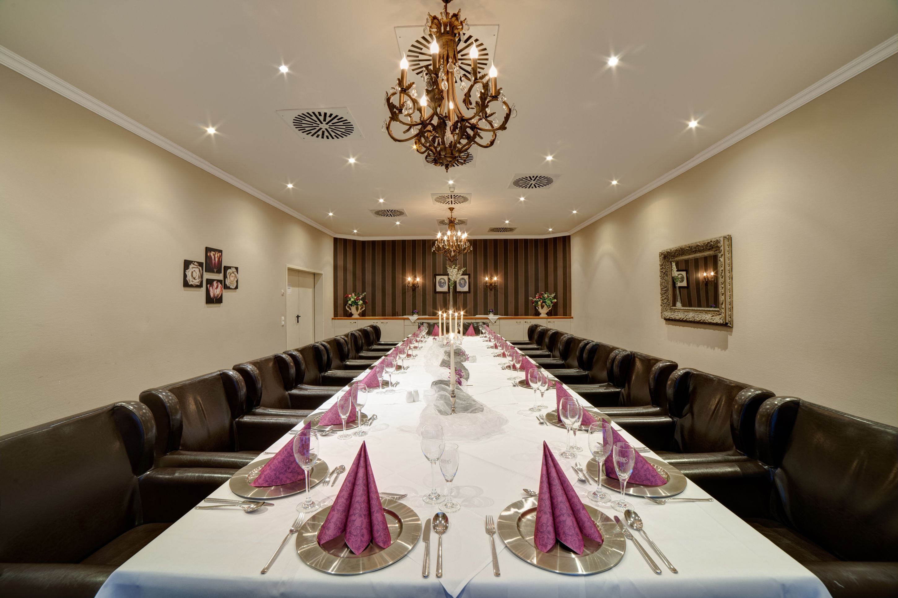 A long dining table set for a formal meal with a chandelier above, leather booth seating, and framed artwork on the walls.
