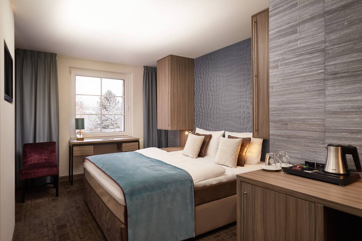 Une chambre d’hôtel équipée d’un lit, d’un bureau, d’une chaise, d’une machine à café et d’une fenêtre donnant sur la verdure.