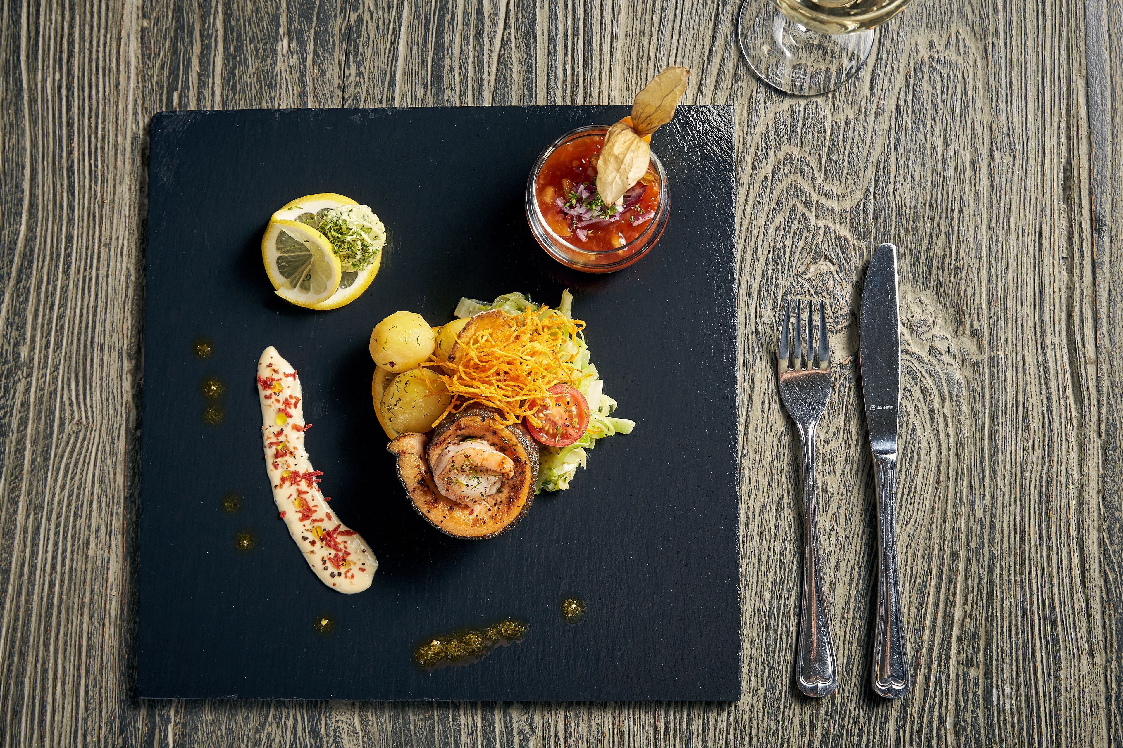 Une planche de service en ardoise noire présente une variété d’aliments, notamment un légume grillé, un morceau de viande et un petit bol de sauce, le tout posé sur une table en bois.