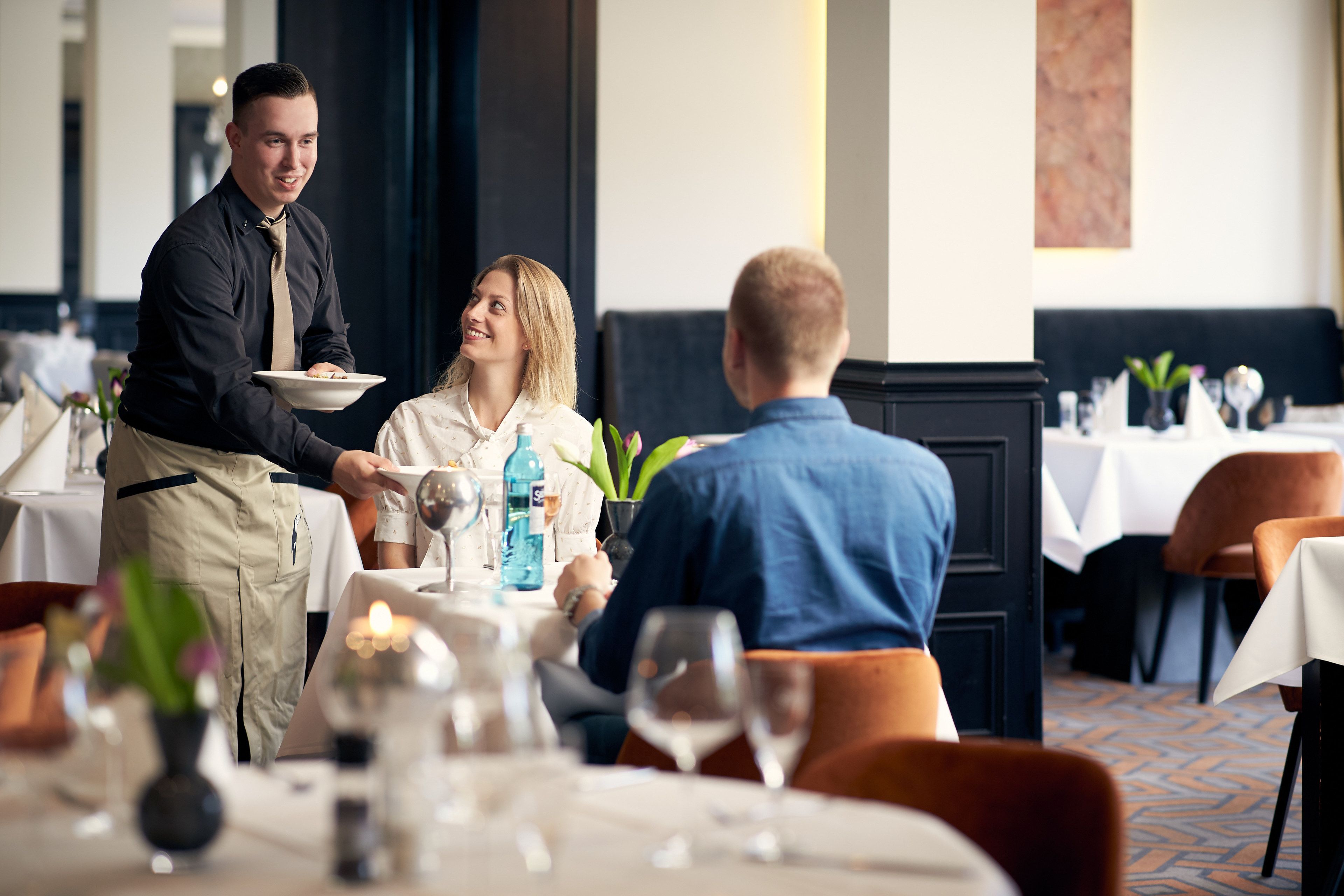 Un ambiente de restaurante con un camarero atendiendo a un cliente. Las mesas están dispuestas con copas de vino y una botella de agua.