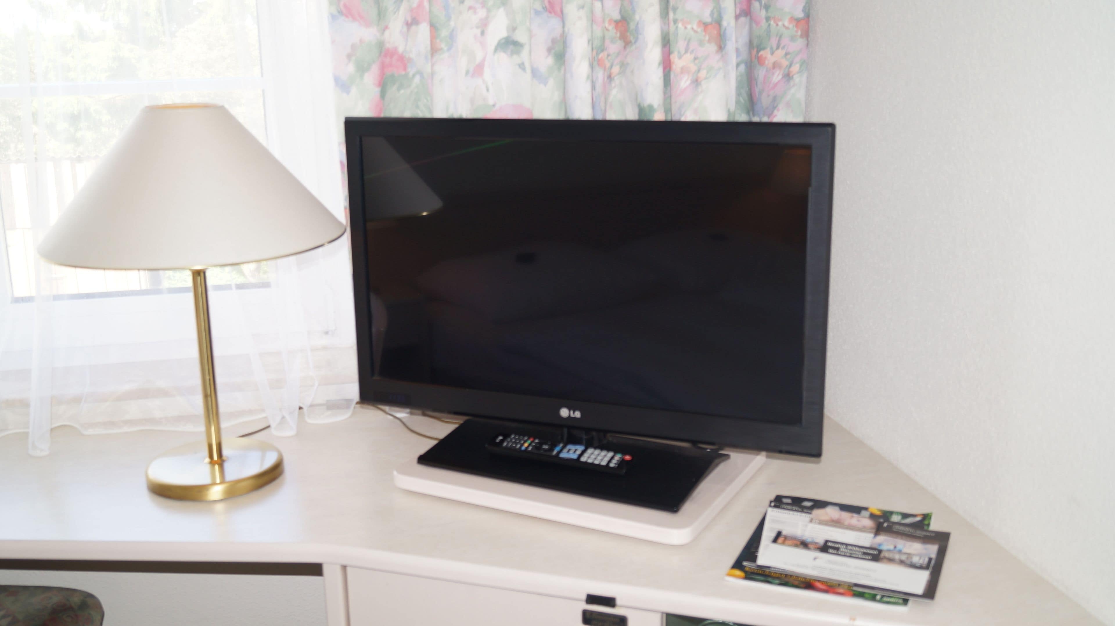 A flat-screen TV on a white desk with a lamp and a brochure.