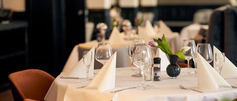 A dining table set with white tablecloths, clear glassware, and a small vase with a flower.