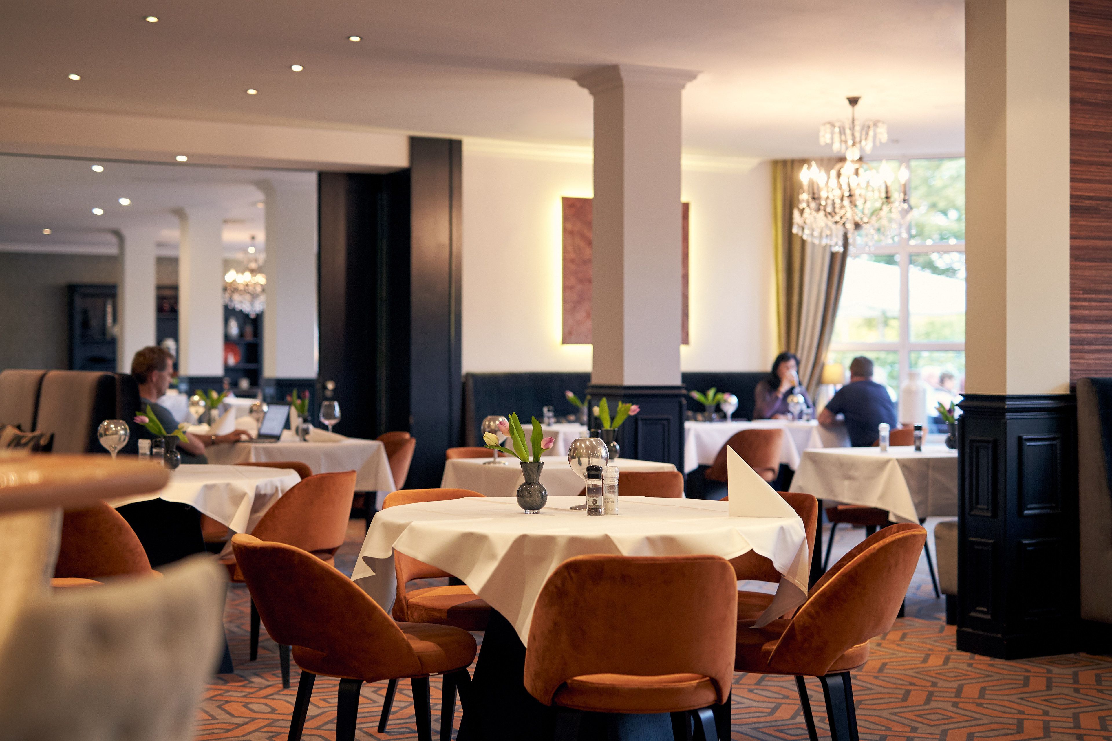 A restaurant with round tables, orange chairs, and a chandelier.