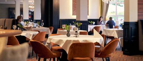 A restaurant with round tables, orange chairs, and a chandelier.
