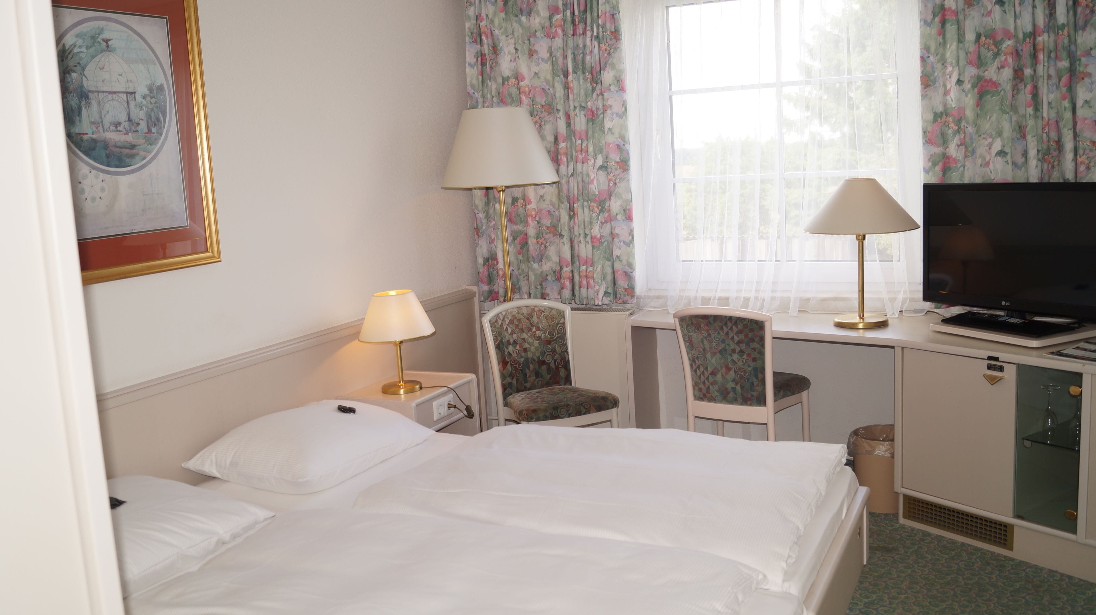 A hotel room with a bed, two chairs, a desk with a TV, and a window with curtains.