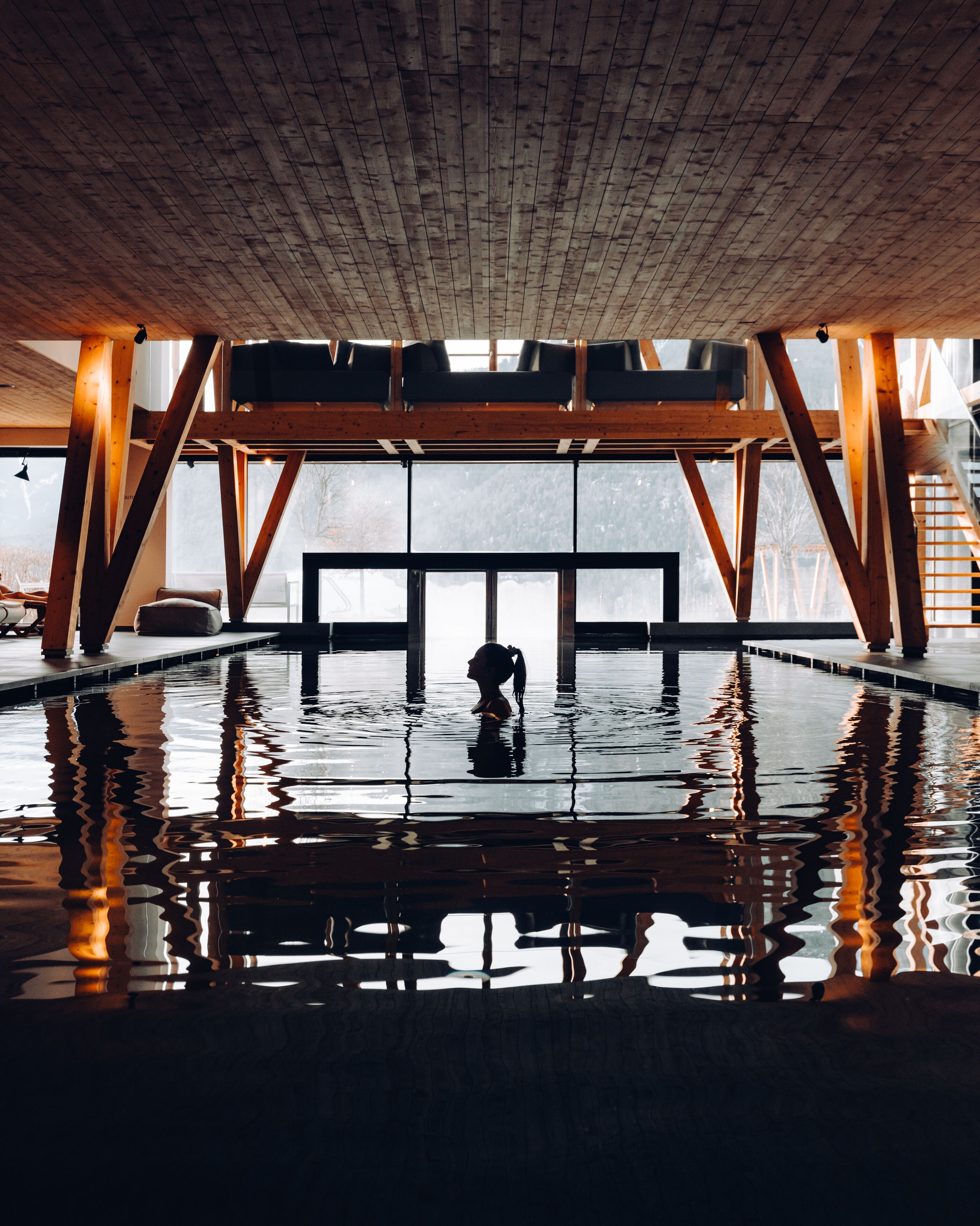 A large indoor swimming pool with wooden beams and a view of a snowy landscape through large windows.