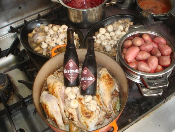 Una cocina con una olla que contiene pollo y champiñones, una sartén con papas, y un bol de papas rojas. Hay dos botellas de cerveza Westmalle Trappist Dubbel.
