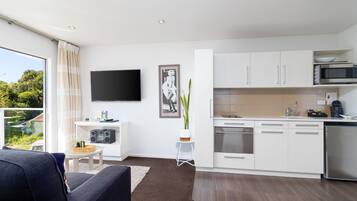 A modern kitchen with white cabinets, a microwave, and a dishwasher. There is a dining table with chairs, a small side table, and a potted plant. A flat-screen TV is mounted on the wall.