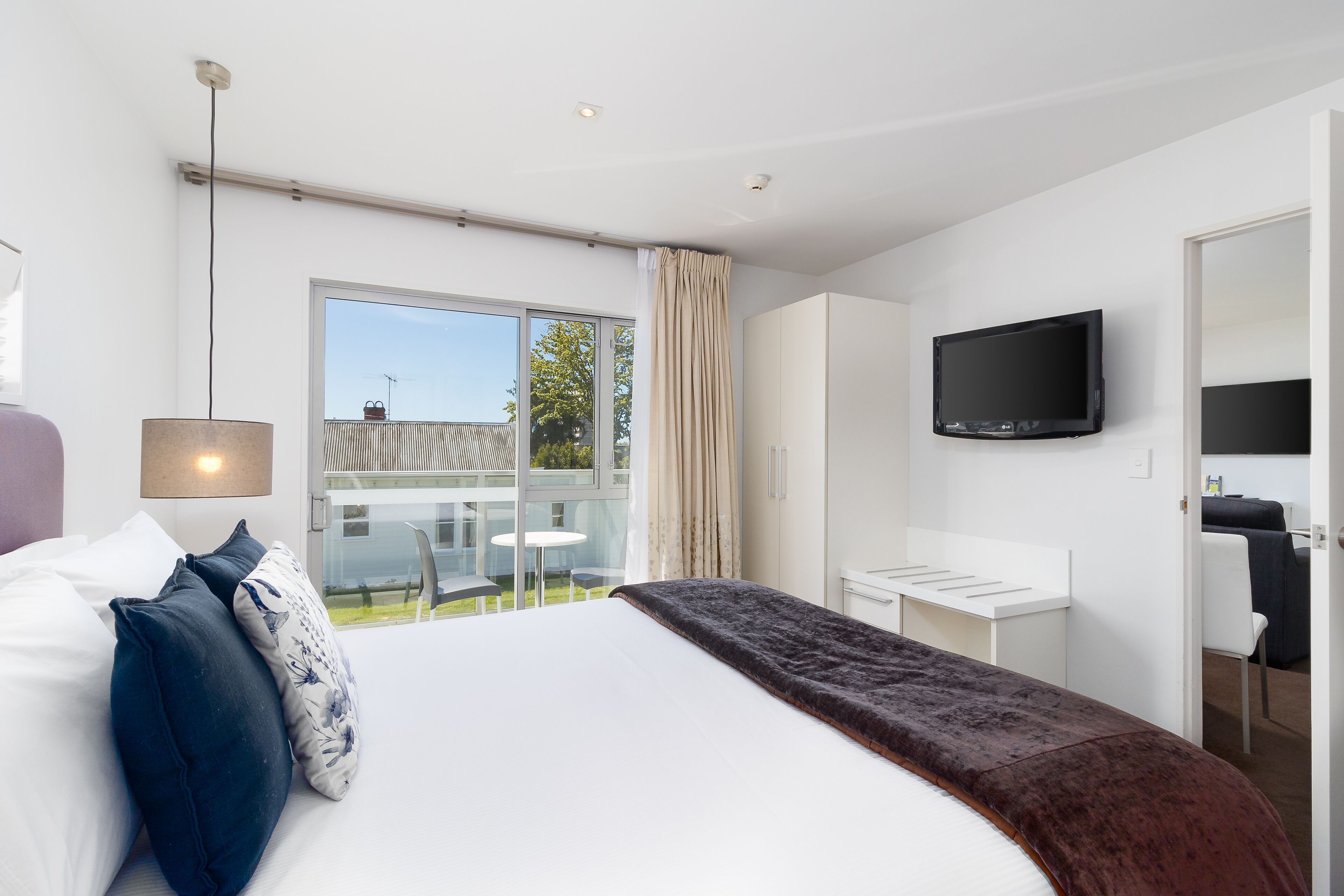 A modern hotel room with a large bed, a TV mounted on the wall, a desk, and a balcony with a glass door.