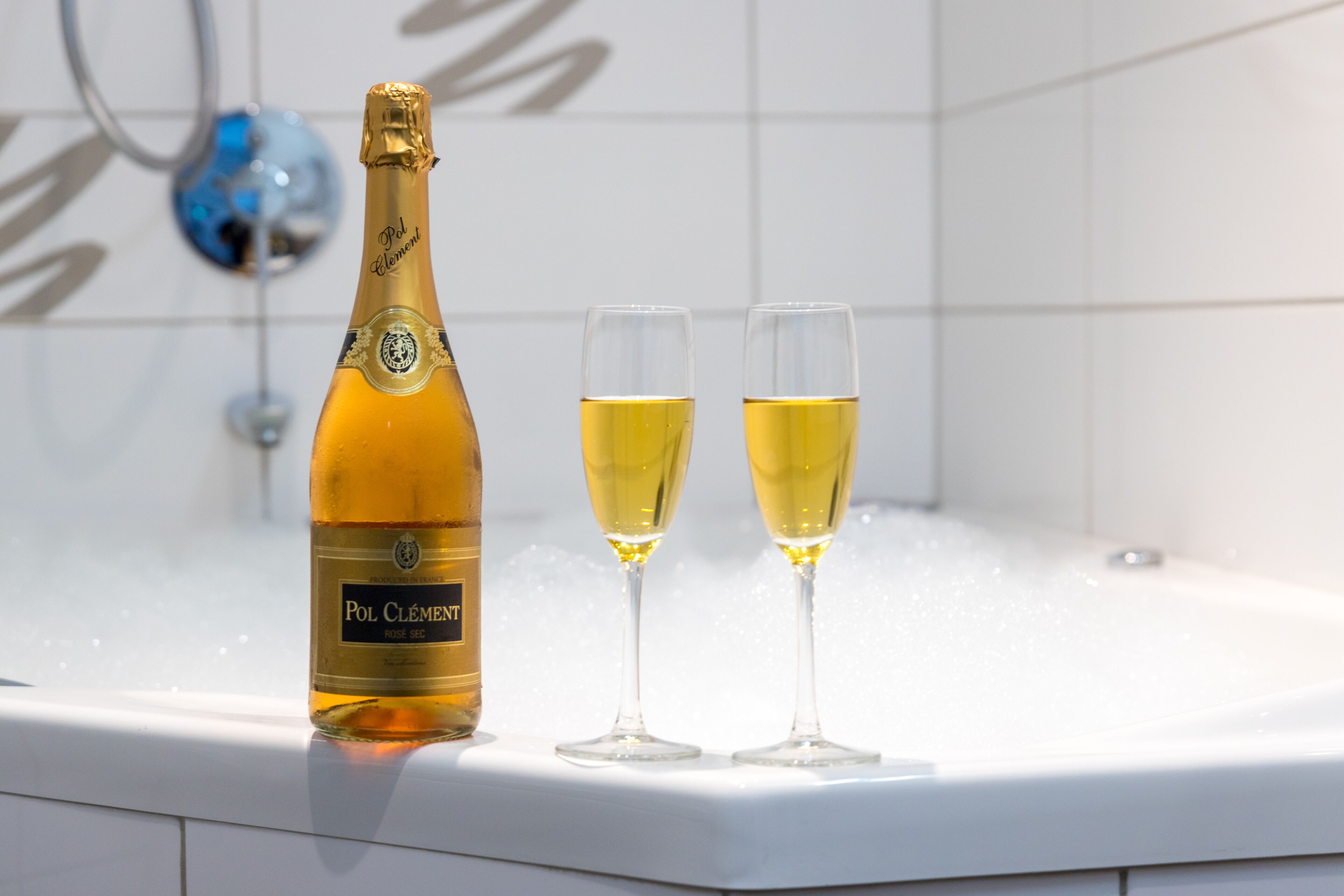A bottle of Pol Clément champagne and two glasses on a white bathtub.