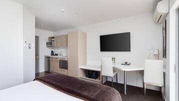 A modern hotel room with a bed, a desk, a chair, a TV, a kitchenette, and an air conditioning unit.