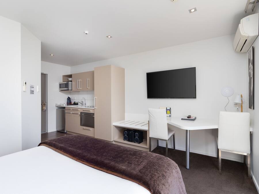 A modern hotel room with a bed, a desk, a chair, a TV, a kitchenette, and an air conditioning unit.