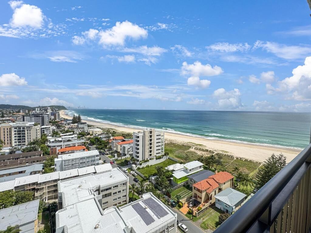 A coastal cityscape with a beach view, featuring residential buildings, a clear sky, and a calm sea.