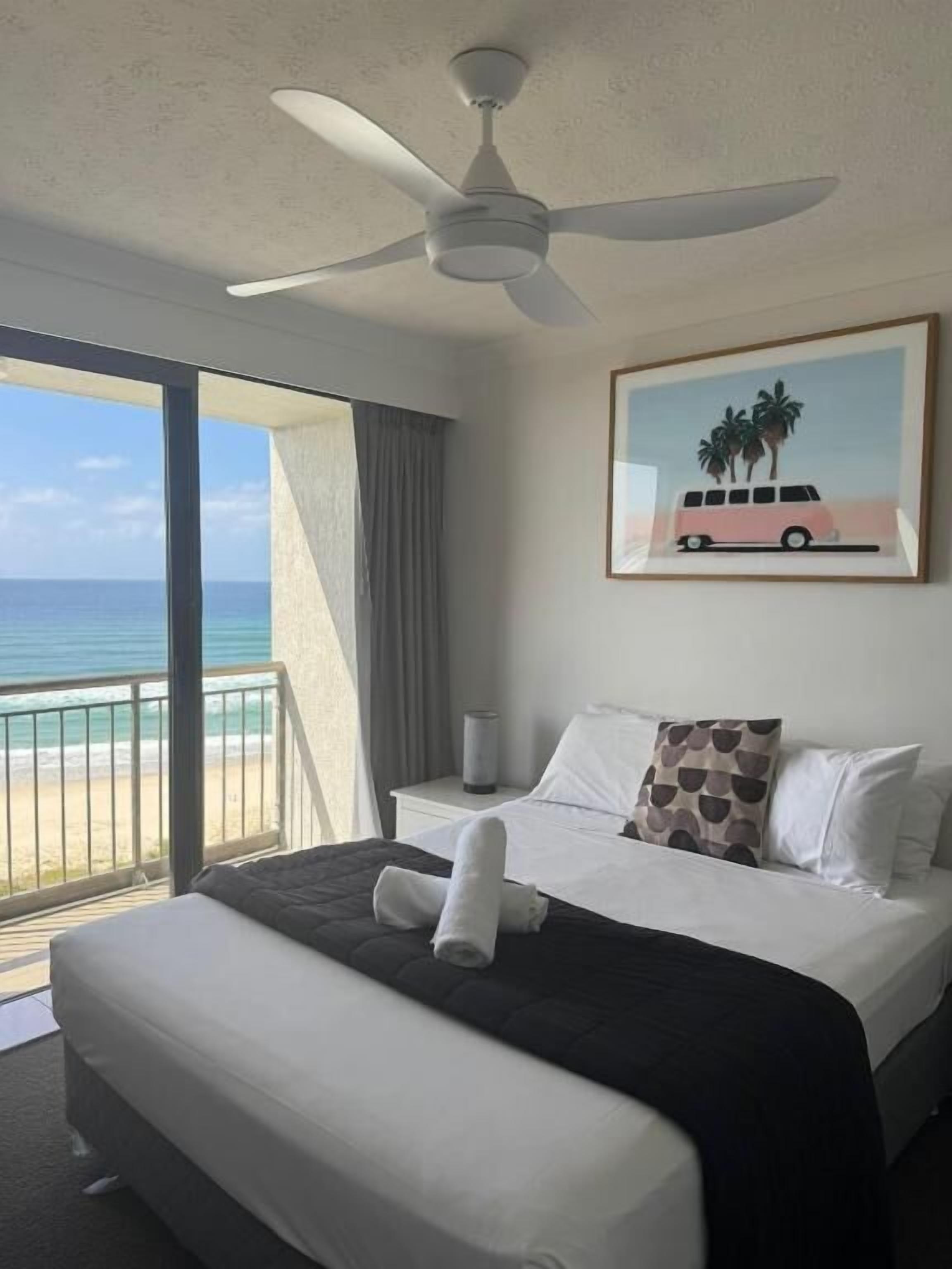 A hotel room with a bed, a ceiling fan, a painting of a van and palm trees, and a view of the ocean.