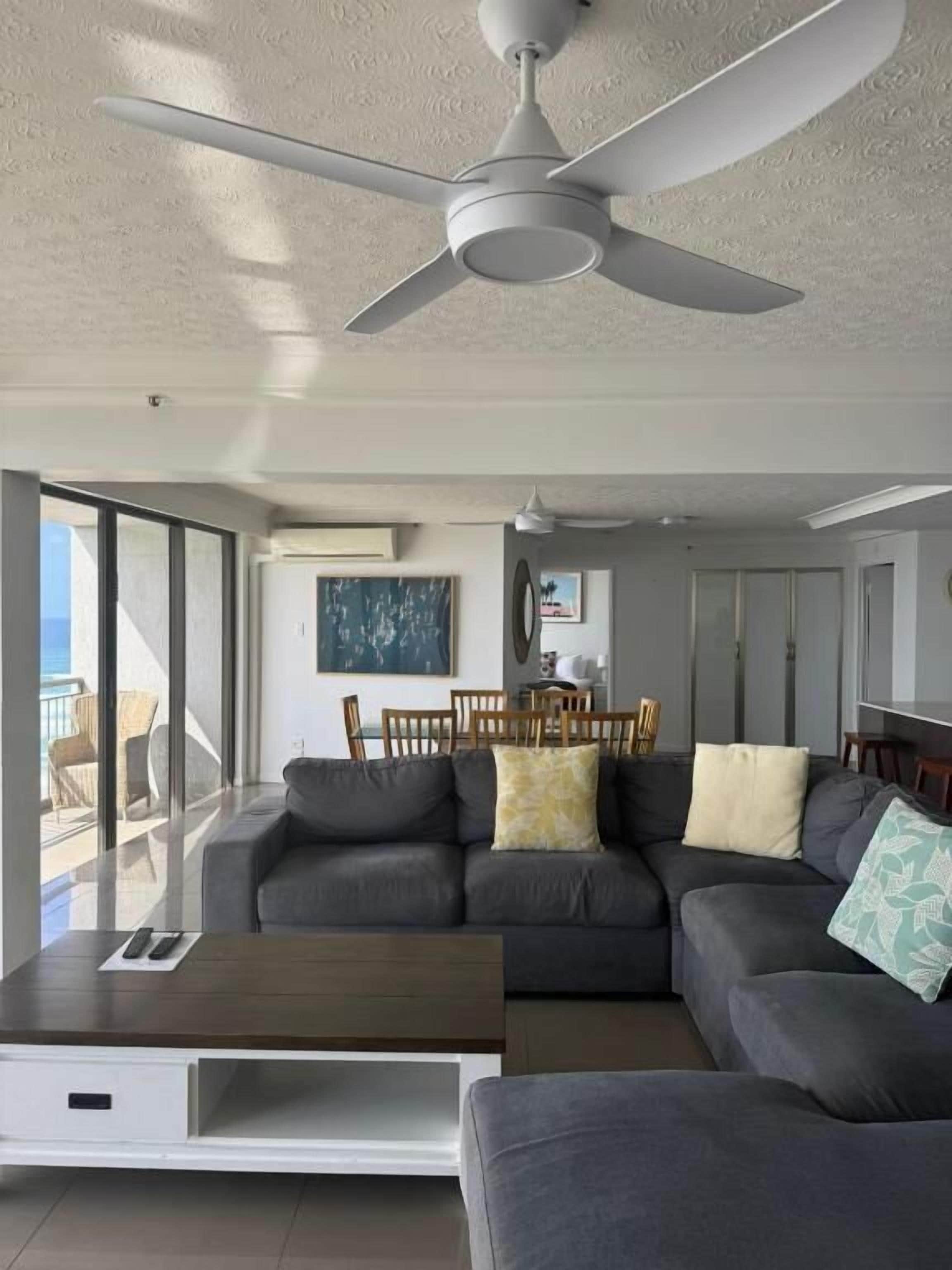 Un salon moderne avec un canapé gris, une table basse et un ventilateur de plafond. On y trouve également un coin repas avec une table et des chaises, ainsi qu’une grande fenêtre offrant une vue dégagée.