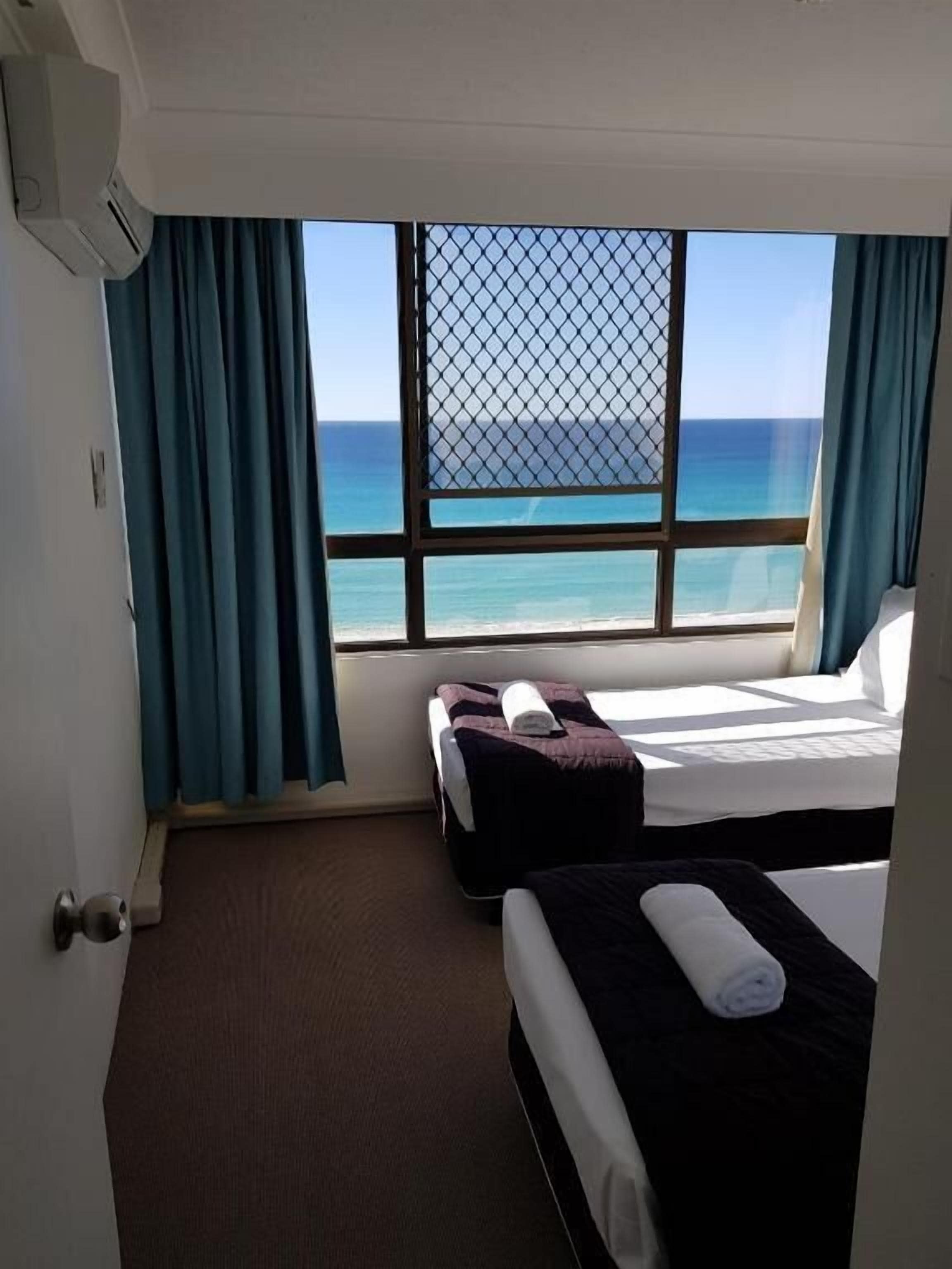 A hotel room with two beds, a large window with a view of the sea, and a ceiling fan.