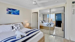 Une chambre d’hôtel moderne avec un grand lit, un ventilateur de plafond, un tableau au mur et une vue sur l’extérieur, visible à travers une porte vitrée.