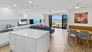 Une cuisine moderne avec des meubles blancs et un grand îlot central, un coin repas avec une table et des chaises en bois, et un salon avec un canapé gris et une grande fenêtre donnant sur un paysage urbain.