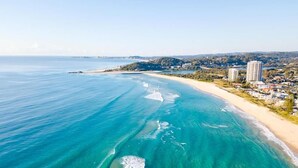 Vue aérienne d’une zone côtière avec des eaux turquoise limpides, une plage de sable et des immeubles résidentiels le long de la côte.
