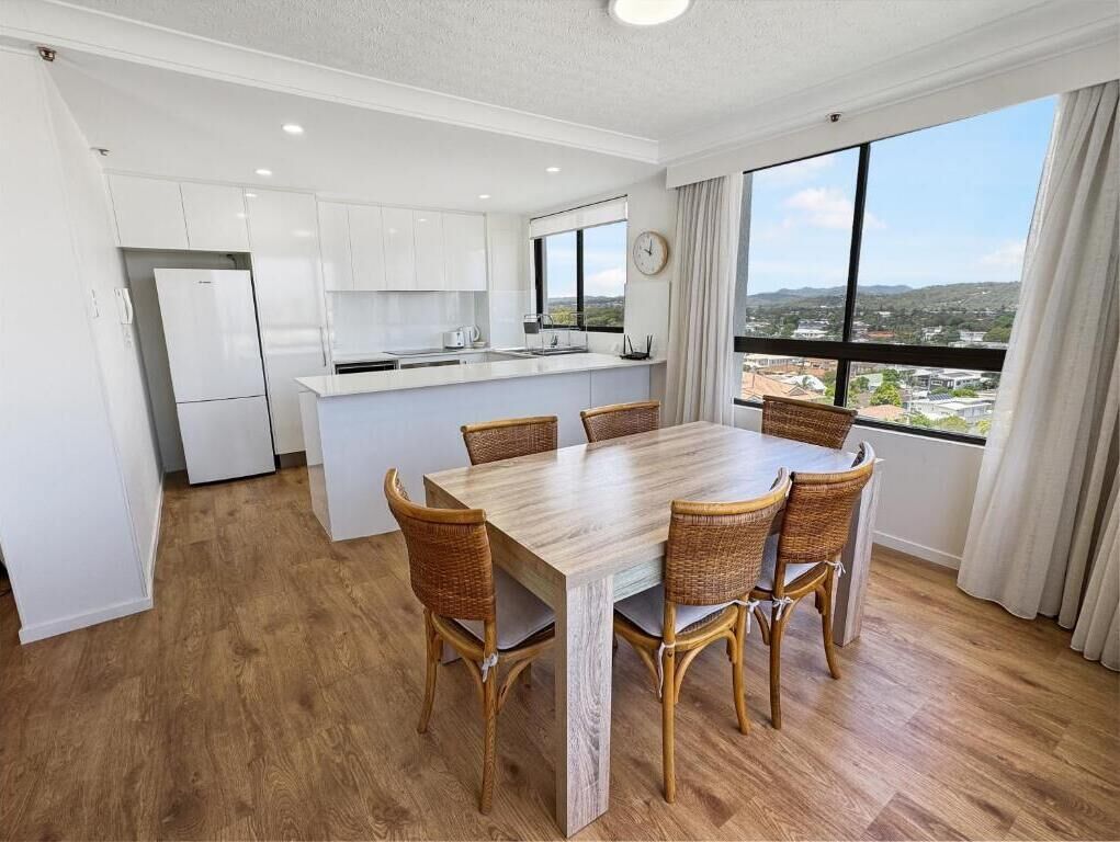 A modern kitchen with white cabinets and a central island, a dining area with a wooden table and wicker chairs, and large windows offering a city view.