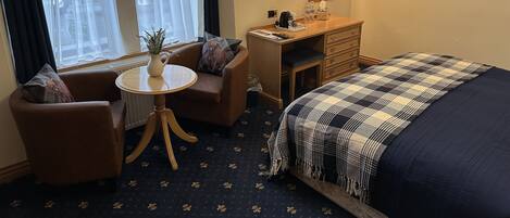 A hotel room with a bed, a desk, a chair, a small table with a plant, a TV, and a window with curtains.