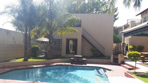 Une piscine avec un bord carrelé, un patio couvert avec des chaises et un escalier menant à un deuxième étage.