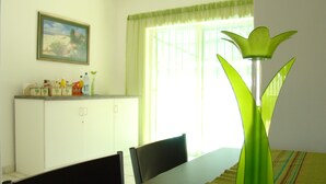 A dining area with a long table, chairs, a cabinet with kitchen items, a painting on the wall, and a window with curtains.