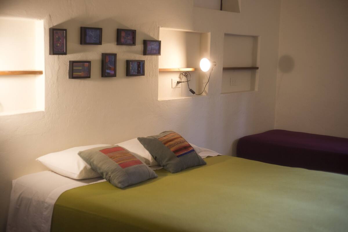Un dormitorio con una cama, almohadas, una lámpara de noche y cuadros en la pared.