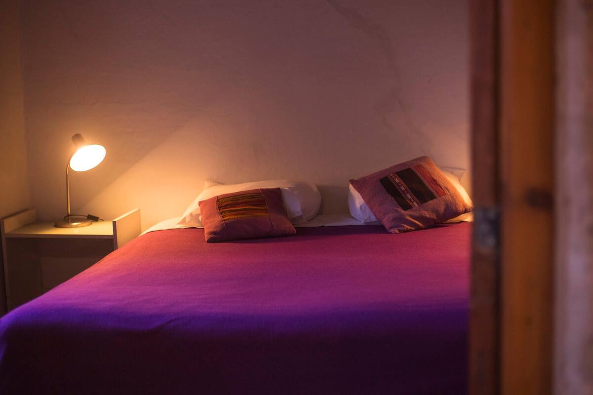 Una cama con colcha morada y dos almohadas, una mesita de noche con lámpara y un marco de puerta de madera.