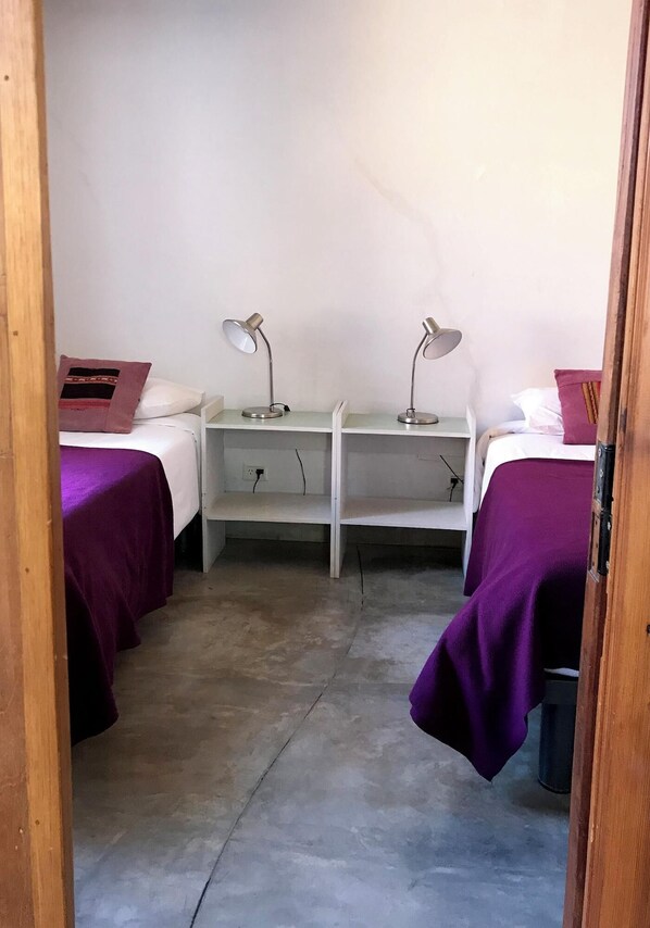 Um quarto pequeno e estreito com duas camas, cada uma com um cobre-cama roxo e uma almofada. Há duas mesinhas de cabeceira com abajures, e uma estante branca no centro.