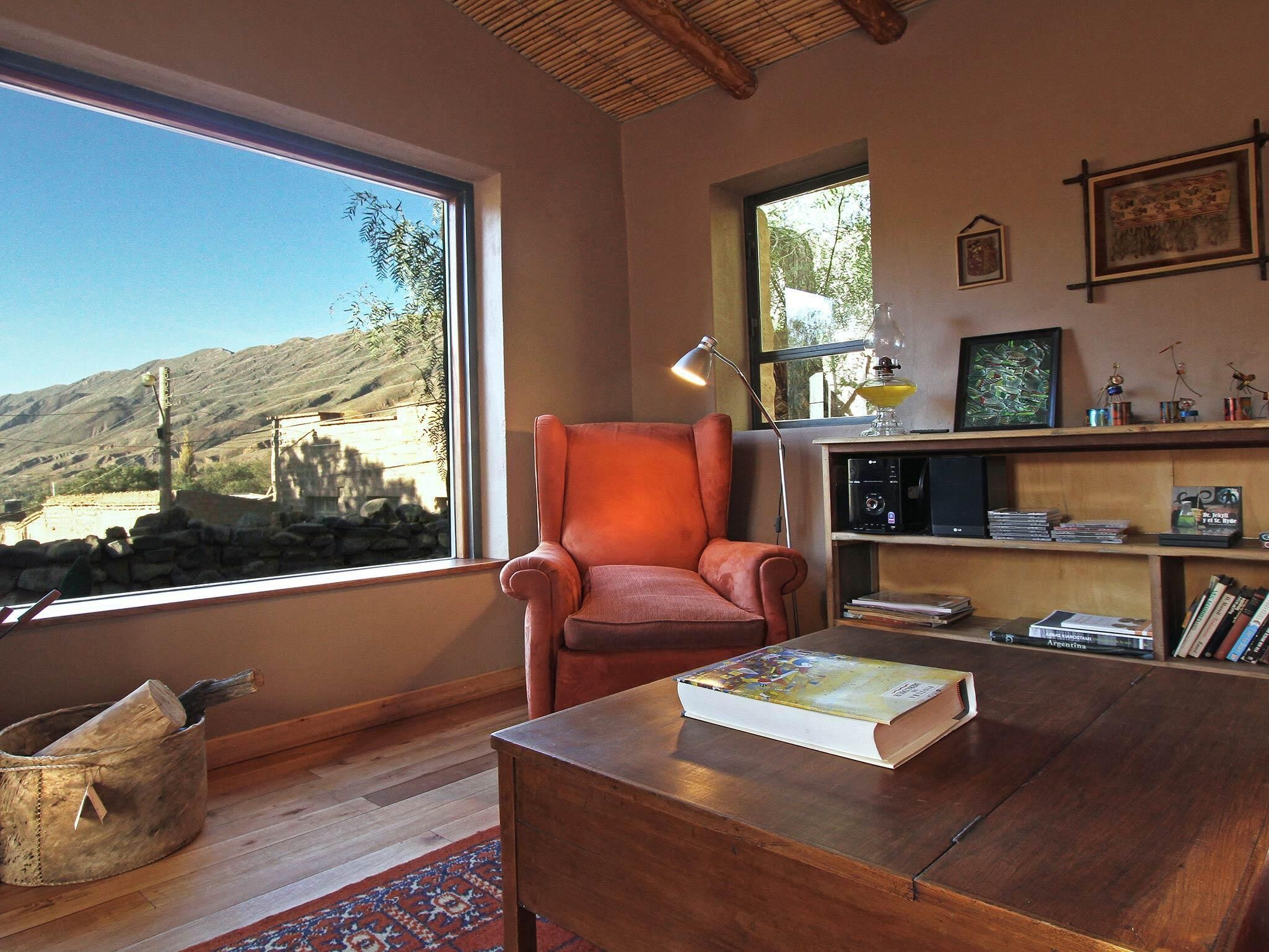 A cozy room with a large window, a wooden table, a red armchair, and a bookshelf filled with books and decorative items.