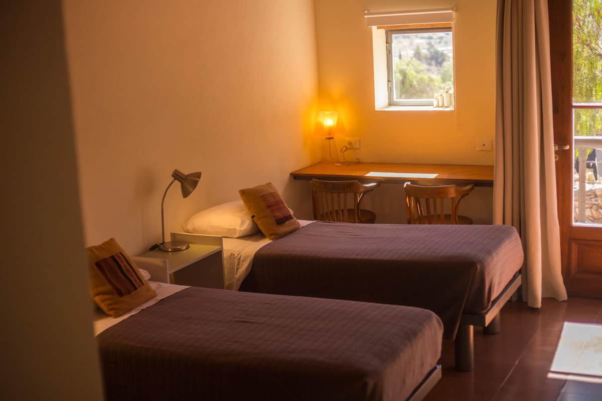 Una habitación con dos camas, un escritorio, una silla, una lámpara y una ventana con cortinas.