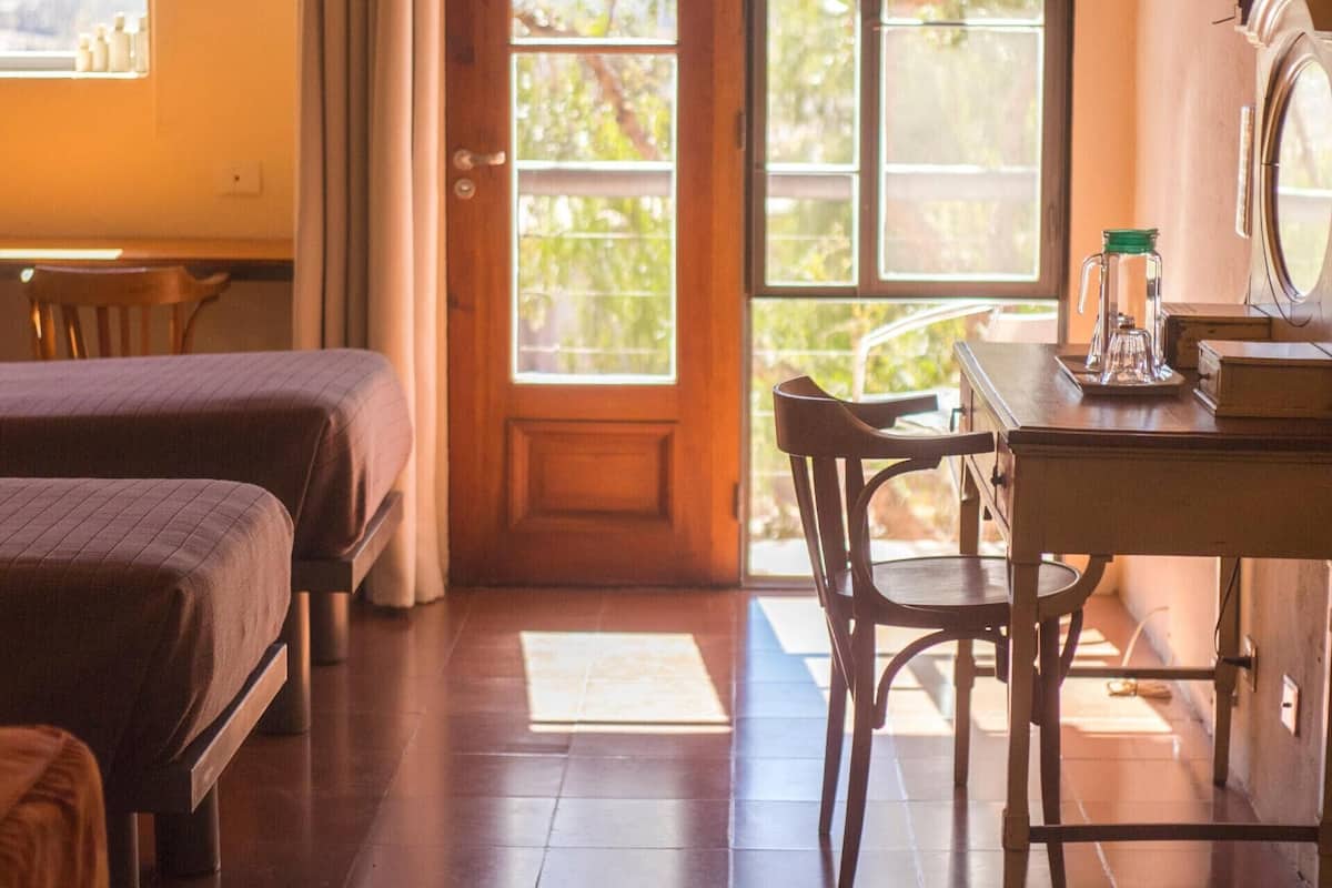 Una habitación con dos camas, una mesa de madera con una silla, una ventana con cortinas y una lámpara de pared.