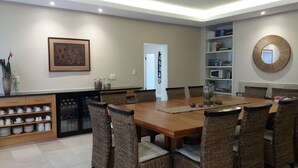 A dining area with a wooden table and wicker chairs, a kitchen area with a dishwasher, and a bookshelf with books and decorative items. - La Roche Guest House (Cape Town)