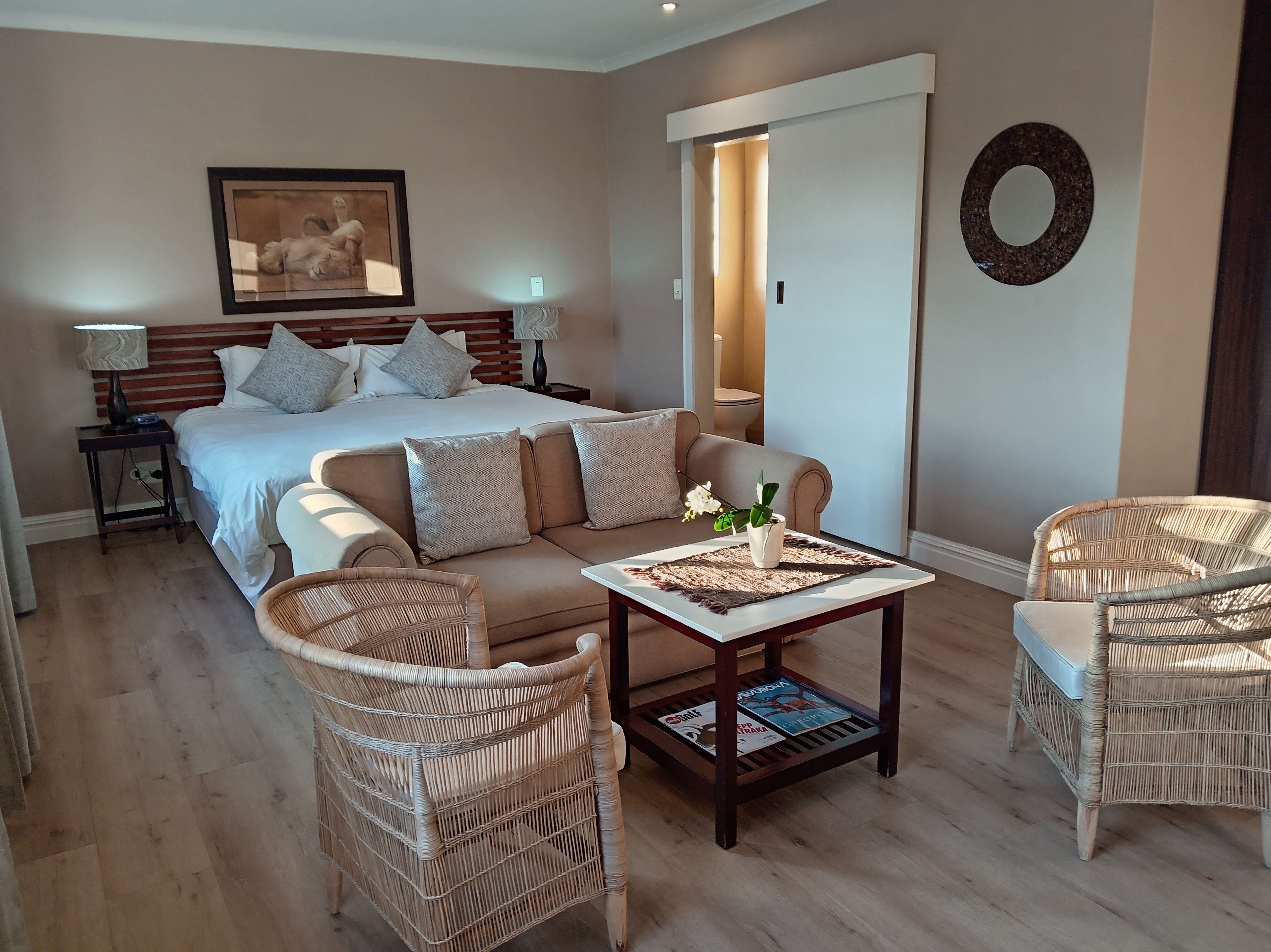 A hotel room with a bed, a sofa, two wicker chairs, a coffee table with magazines, a framed picture on the wall, and a door leading to another room.