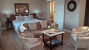 Une chambre d’hôtel comprenant un lit, un canapé, deux fauteuils en osier, une table basse avec des magazines, un tableau encadré au mur et une porte donnant sur une autre pièce.