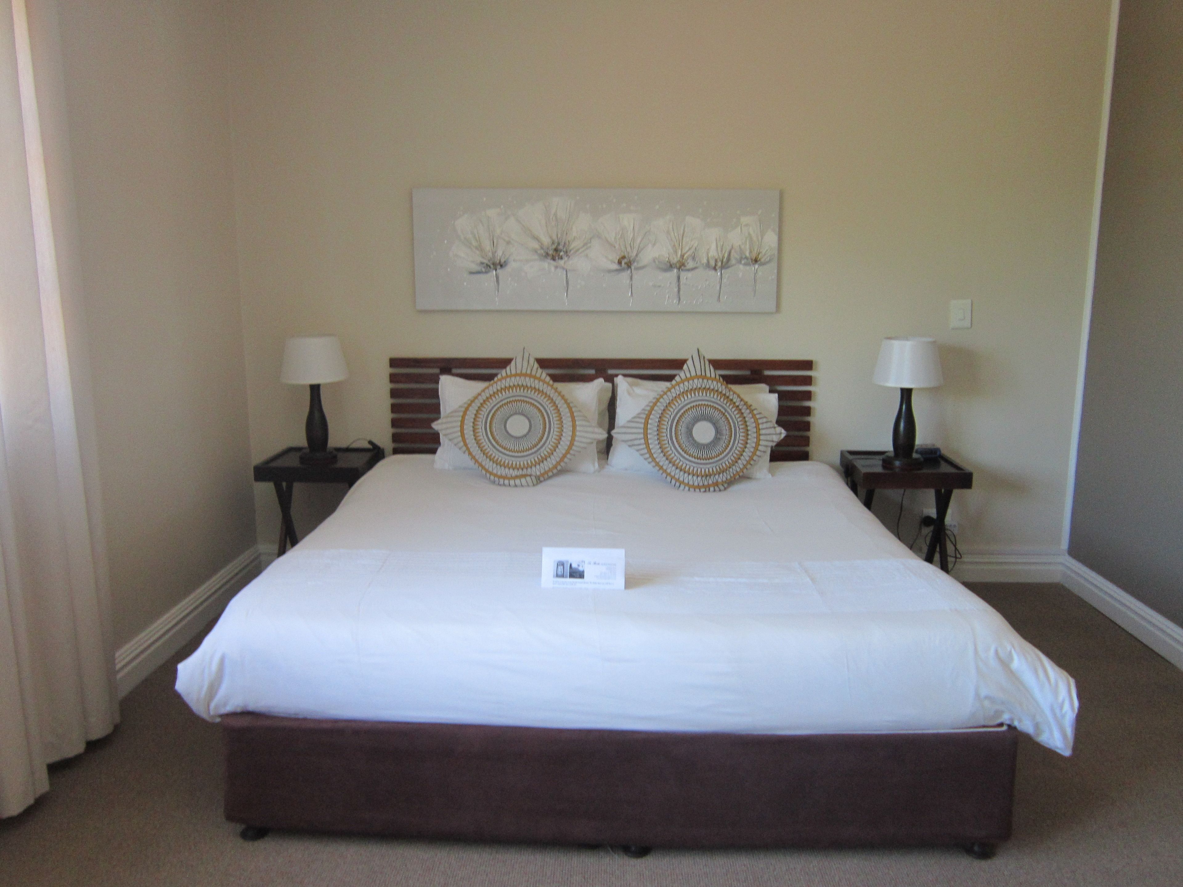 Quarto de hotel com uma cama grande, duas mesas de cabeceira com abajures, um quadro na parede e piso coberto por carpete.