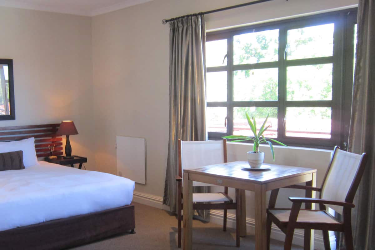 Una habitación de hotel con una cama grande, una mesita de noche con lámparas, un cuadro enmarcado en la pared, una ventana con cortinas y una pequeña mesa con una planta.