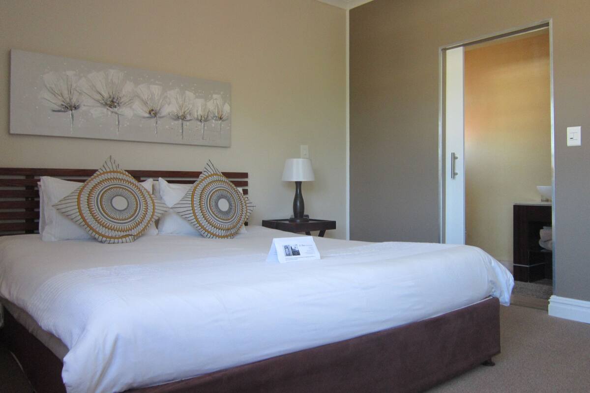 Habitación de hotel con una cama grande, una mesita de noche, una lámpara y una puerta que da a otra habitación.