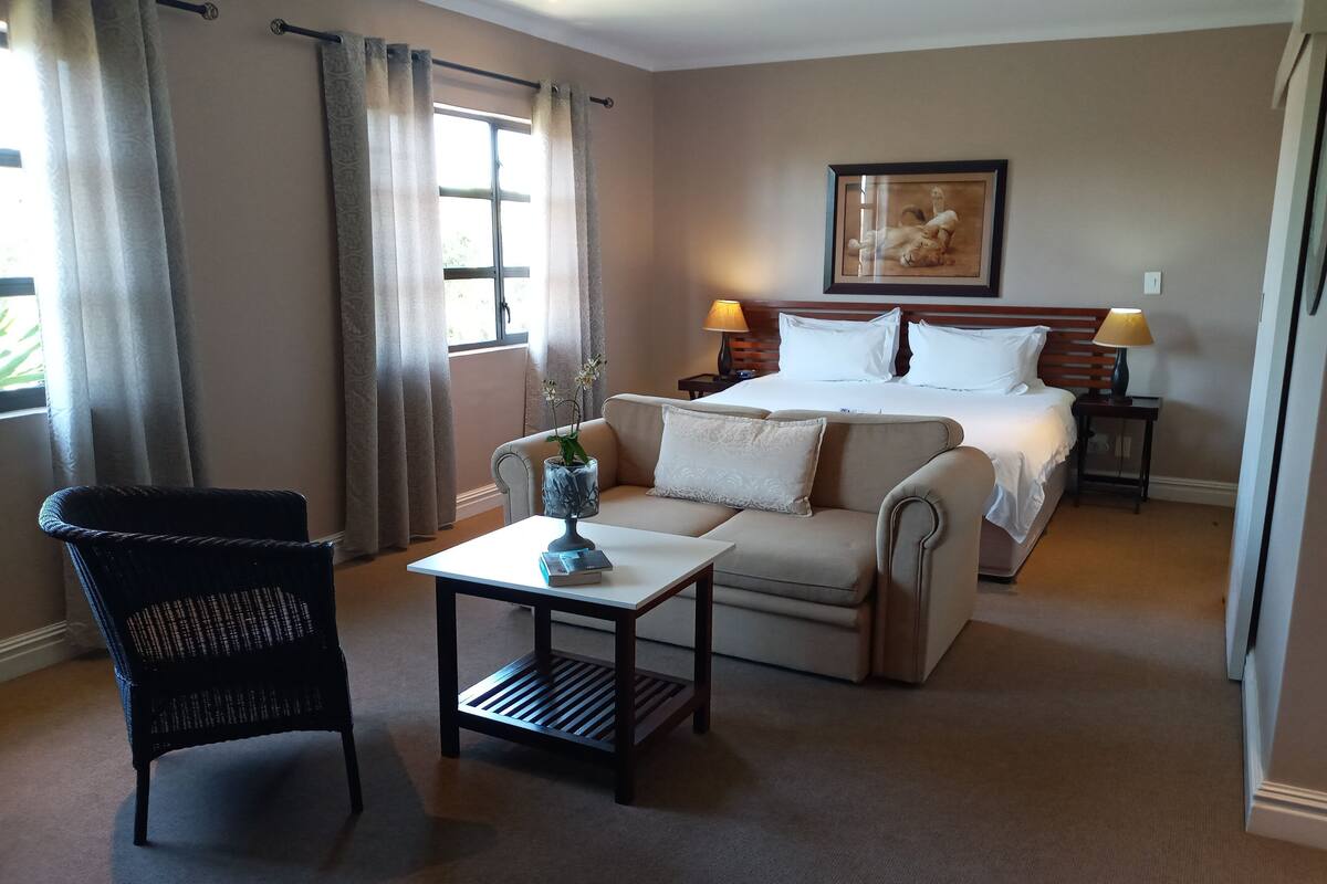 Habitación de hotel con cama, dos mesitas de noche con lámparas, un sofá, una silla y una pequeña mesa con un jarrón y un libro.