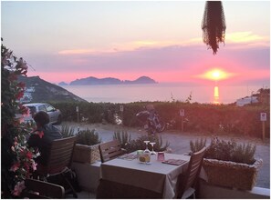 A table set outdoors with a view of the sunset over the sea, a parked car, and a motorcycle.