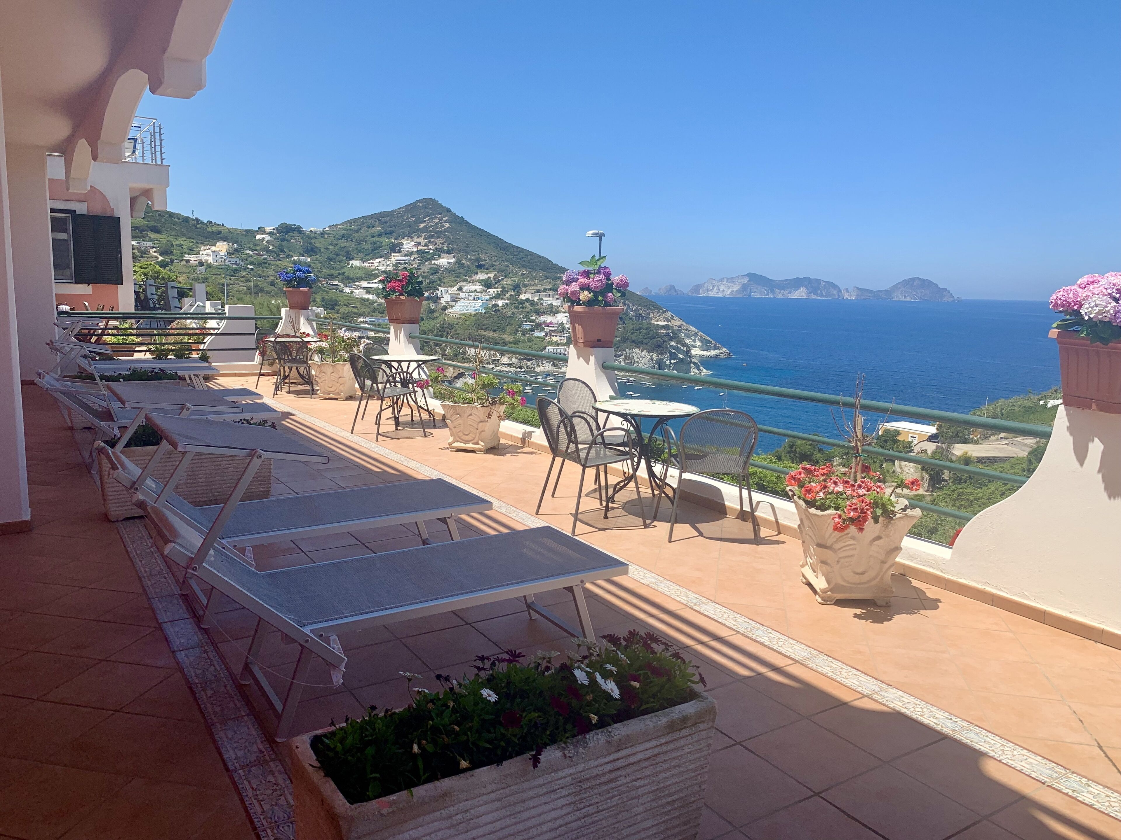 Une terrasse dotée de chaises longues et de tables, agrémentée de plantes en pot, et offrant une vue sur la mer et les collines.