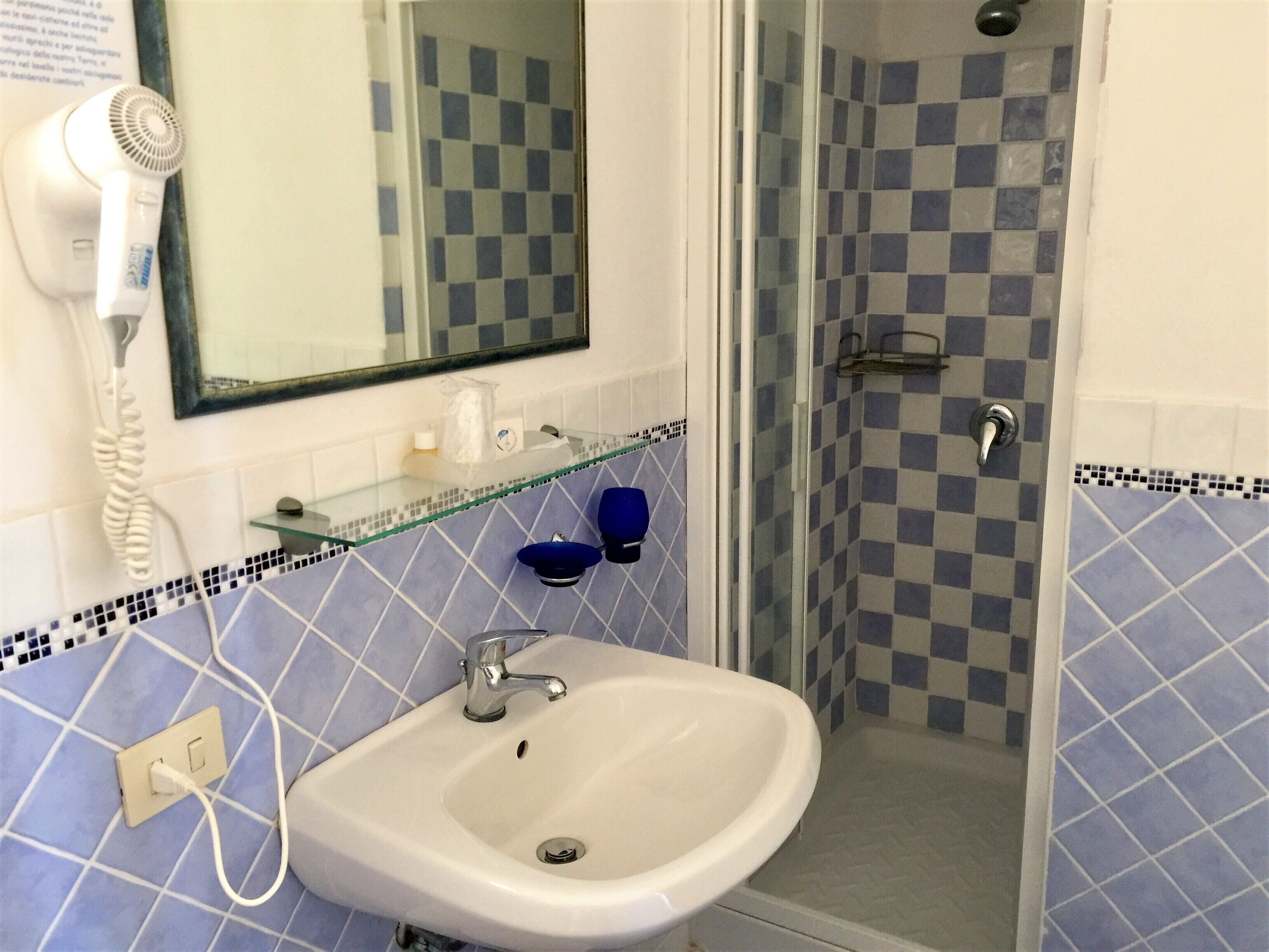 Une salle de bain avec un lavabo blanc, une étagère en verre, une douche bleue et un sèche-cheveux fixé au mur.