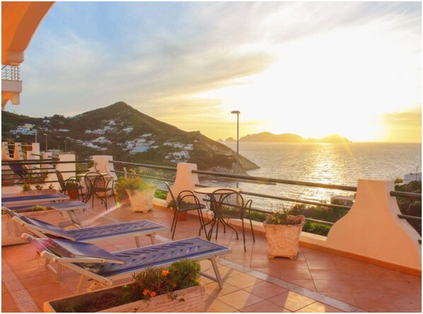 A terrace with sun loungers, potted plants, and a view of the sea and a mountain at sunset.